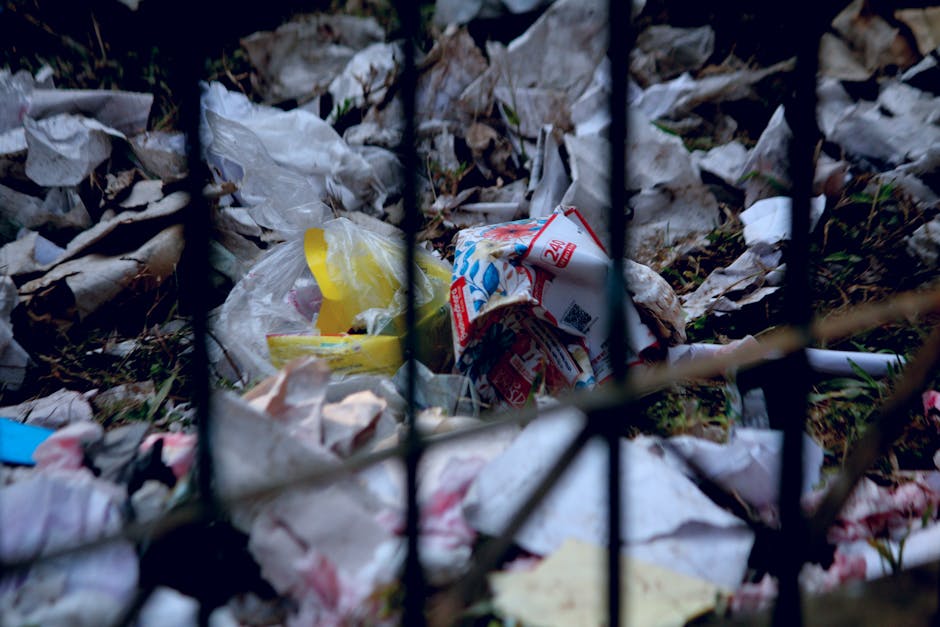 A brown vintage-style sofa with ornate wooden carved details on the arms and backrest is positioned in front of a large wall of tightly packed, overflowing rubbish bags and loose waste materials, incl