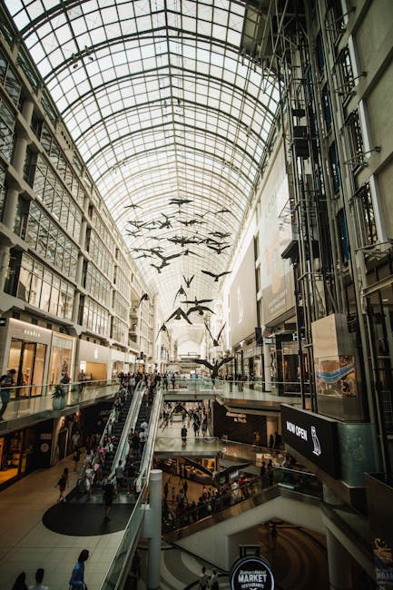 Interior of a modern shopping mall with a high glass vaulted ceiling allowing natural light to illuminate the space. The ceiling features a large installation of multiple black bird-shaped sculptures
