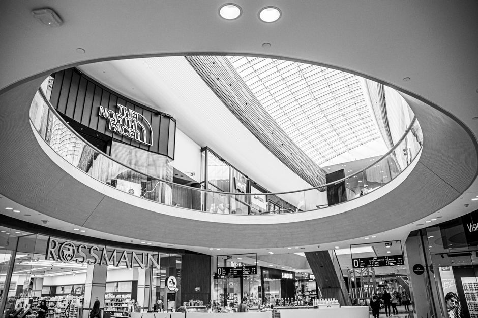 Interior of a modern shopping mall with a high glass vaulted ceiling allowing natural light to illuminate the space. The ceiling features a large installation of multiple black bird-shaped sculptures