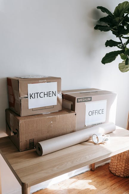 A close-up view of a person's hand placing or removing bubble wrap inside a cardboard box labeled 'KITCHEN' for a home relocation. The box, made of brown cardboard, is set on a surface near a door or