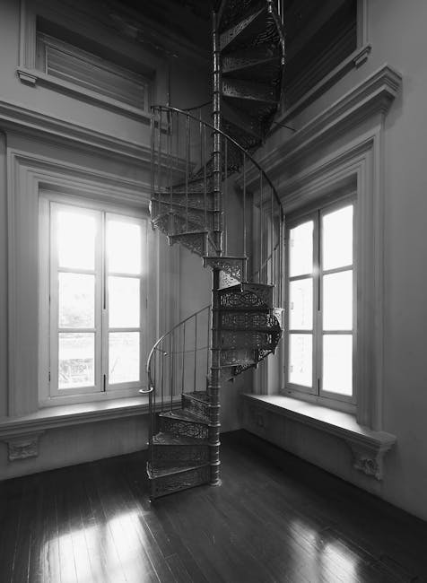 A narrow outdoor staircase with gray steps ascending between high, beige-colored walls. On the left side, there are visible electrical conduits, boxes, and meters attached to the wall, while the right